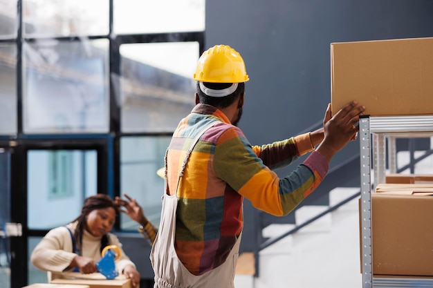 Gestionnaire d'entrepôt debout sur une échelle tout en atteignant une boîte en carton. Chargeur d'entrepôt afro-américain portant un casque de sécurité et une étagère de déchargement de vêtements de travail avec des colis lourds