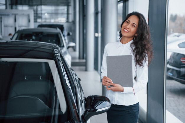 Gestionnaire féminine aux cheveux bouclés se tient près de la voiture dans le salon automobile