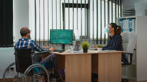 Gestionnaire handicapé venant avec un fauteuil roulant sur le lieu de travail avec un masque de protection travaillant dans un nouveau bureau d'affaires normal. Indépendant immobilisé en société financière respectant la distanciation sociale.