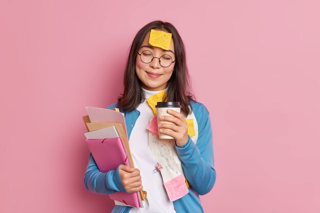 Une gestionnaire satisfaite travaille avec des documents papier a une pause-café ferme les yeux, une note collante avec un graphique dessiné collé au front porte des lunettes rondes.