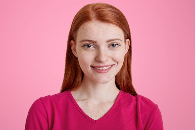 Gingembre femelle avec un sourire agréable, a la peau de rousseur, étant heureux d'être photographié, habille en pull rose