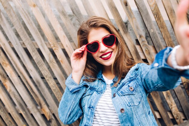 Glamour fille souriante portant des lunettes coeur tenant cadre. Portrait en gros plan de la charmante jeune jolie femme touchant la caméra.