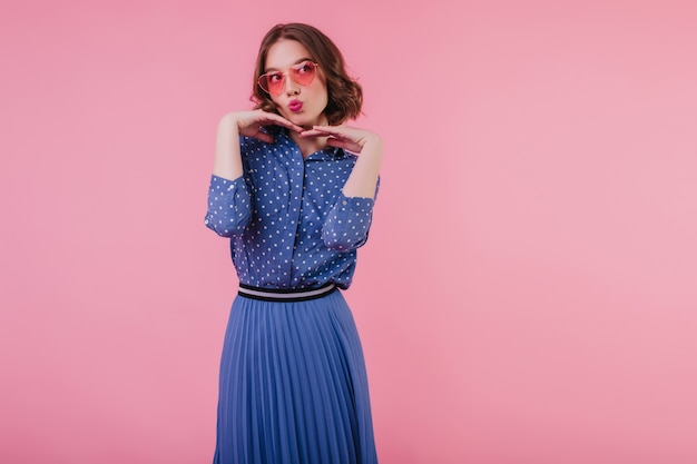 Glamour jeune femme en chemisier bleu drôle posant Photo intérieure d'une adorable fille aux cheveux courts en lunettes de soleil roses.