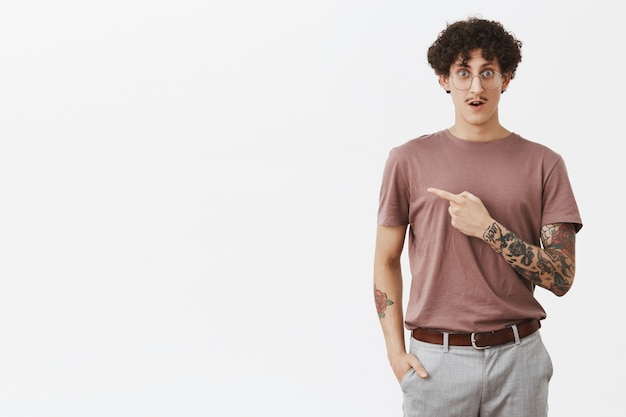 Gosh regarde cette nana. Impressionné et stupéfait beau mec urbain élégant avec une coiffure frisée et un bras tatoué ouvrant la bouche par étonnement et curiosité pointant à gauche interrogé sur un mur gris