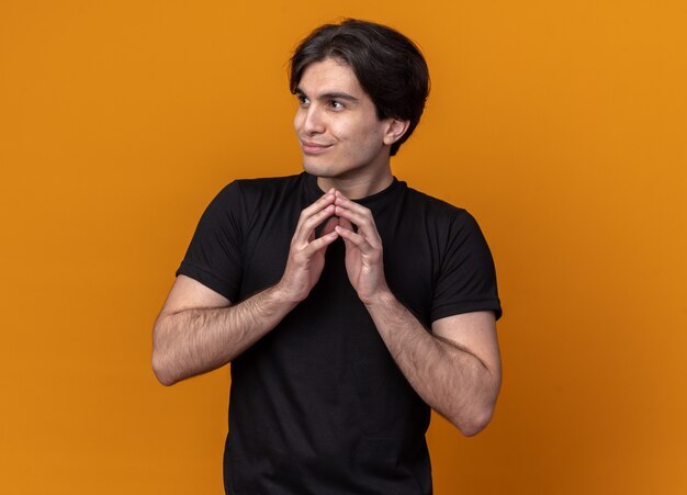 Gourmand regardant le côté jeune beau mec portant un t-shirt noir tenant les mains ensemble isolé sur un mur orange