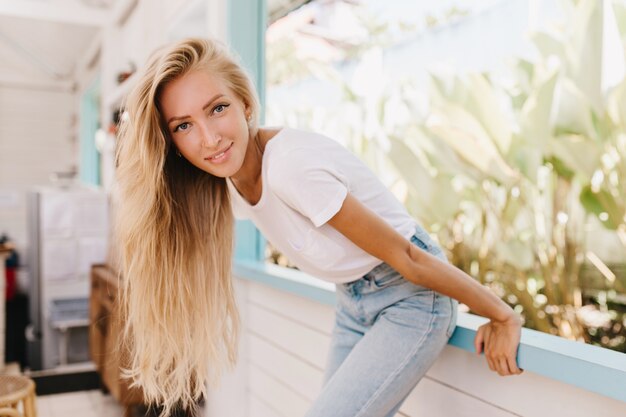 Gracieuse dame aux longs cheveux blonds s'amusant le week-end et posant avec plaisir. Femme caucasienne enthousiaste à la peau bronzée en riant debout près de la fenêtre.