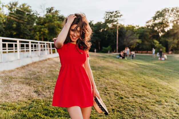 Gracieuse dame caucasienne aux cheveux bruns tenant un chapeau de paille sur la nature Superbe femme brune en robe rouge, passer du temps sur le terrain.