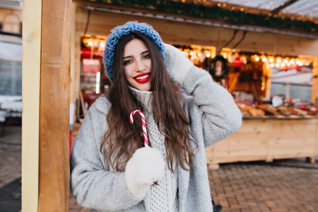 Gracieuse femme aux cheveux noirs avec canne en bonbon souriant. Portrait en plein air d'une superbe fille à la mode dans des mitaines blanches s'amusant sur le marché de Noël.