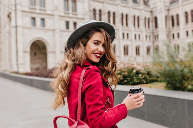 Gracieuse femme blanche au chapeau gris regardant par-dessus l'épaule tout en explorant la vieille partie de la ville