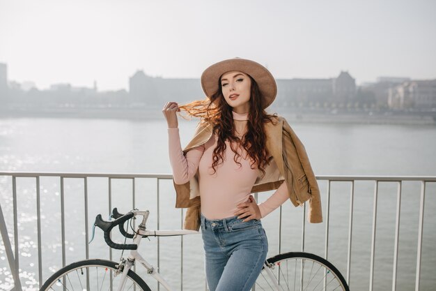 Gracieuse femme mignonne joue avec ses cheveux roux en se tenant debout à côté de vélo