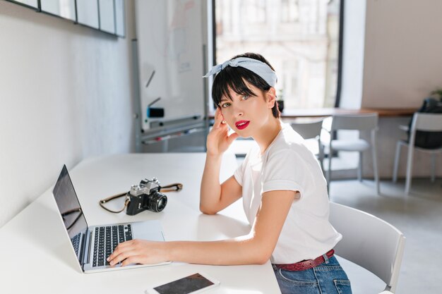Gracieuse femme photographe éditant des photos sur un ordinateur portable assis en grand blanc avec une expression de visage sérieuse