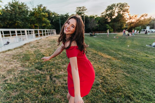 Gracieuse fille aux cheveux noirs exprimant le bonheur. Tir extérieur d'une superbe femme brune en robe rouge s'amusant dans le parc.