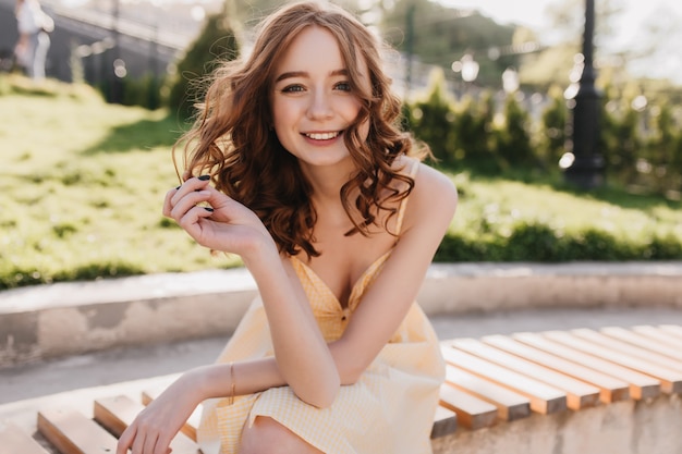 Gracieuse fille de gingembre en robe jaune assis dans le parc. Belle femme frisée aux cheveux roux posant avec un sourire inspiré sur la nature.