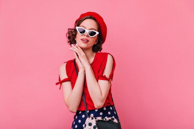 Gracieuse fille pâle en béret français debout. Femme brune optimiste en lunettes de soleil posant.