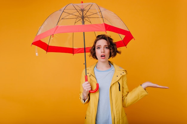 Gracieuse fille porte un élégant manteau d'automne debout sous le parasol. Portrait en studio de modèle féminin caucasien bouleversé posant avec un parapluie sur un mur jaune.