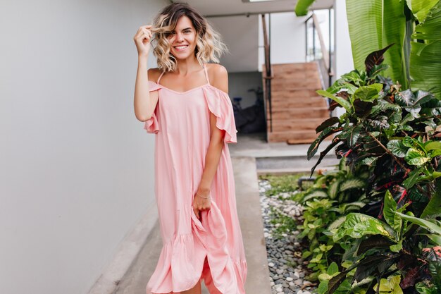 Gracieuse fille souriante en robe longue jouant avec ses cheveux bouclés. Photo extérieure d'une femme bronzée raffinée en tenue rose.