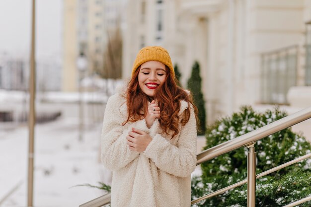 Gracieuse jeune femme au gingembre posant en hiver. Fille caucasienne inspirée en manteau élégant debout dans la rue avec le sourire.