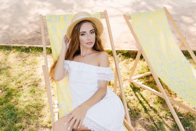 Gracieuse jeune femme regardant avec intérêt tout en étant assise confortablement dans une chaise longue. Fille incroyable en chapeau d'été se faire bronzer dans le jardin en matinée ensoleillée.