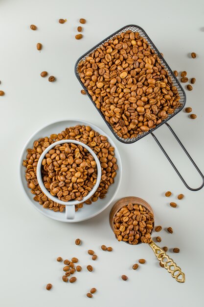 Graines de café aromatisées dans une cafetière, une tasse et une passoire noire sur une surface blanche. mise à plat.