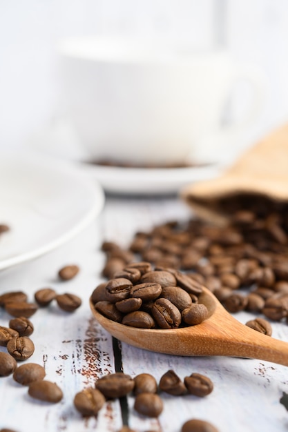 Grains de café sur une cuillère en bois et des sacs de chanvre sur une table en bois blanc.