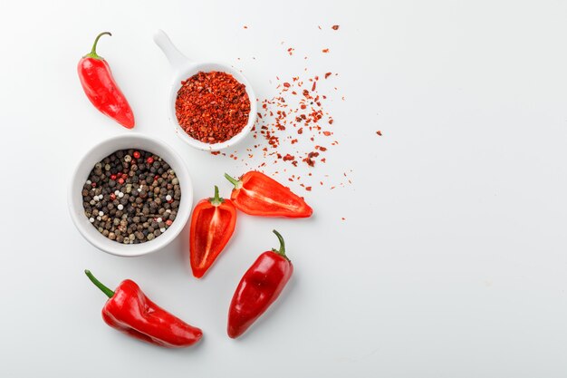 Grains de poivre avec de la poudre de chili en cuillère et poivrons rouges dans une assiette sur mur blanc, vue du dessus.