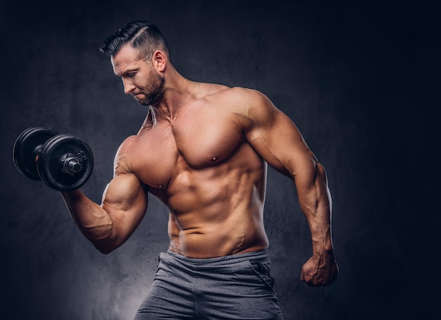 Grand bodybuilder torse nu élégant vêtu d'un short de sport, faisant de l'exercice sur un biceps avec une barre, debout dans un studio. Isolé sur un fond sombre.