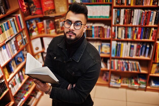 Grand étudiant arabe intelligent portant une veste en jean noir et des lunettes à la bibliothèque avec un livre à la main