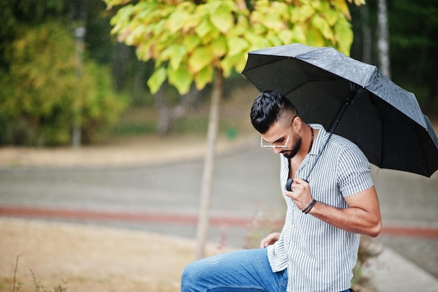 Grand homme à la mode barbe arabe porter sur chemise jeans et lunettes de soleil avec parapluie posé à la pluie sur la place du parc