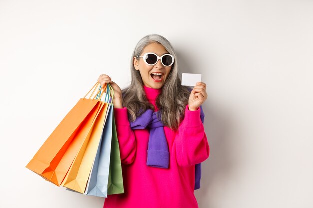 Grand-mère asiatique élégante en lunettes de soleil faisant du shopping en vente de vacances, tenant des sacs en papier et une carte de crédit en plastique, debout sur fond blanc.