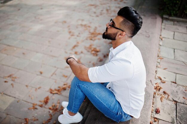 Grand modèle homme arabe élégant en chemise blanche jeans et lunettes de soleil assis dans la rue de la ville Beard attrayant mec arabe au parc