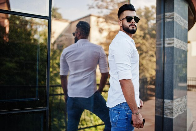 Grand modèle d'homme arabe élégant en chemise blanche, jeans et lunettes de soleil posés dans la rue de la ville Beard attrayant mec arabe contre un bâtiment moderne