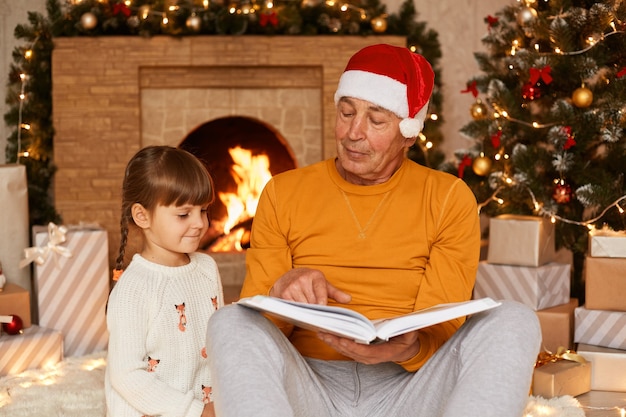 Grand-père portant un pull orange et un chapeau de père noël lisant un livre à sa mignonne petite-fille alors qu'il était assis dans un salon festif près de la cheminée et de l'arbre de noël.