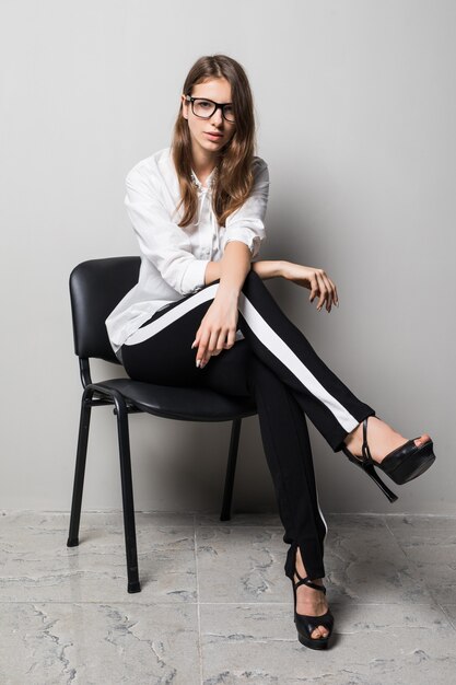 Grande fille brune à lunettes habillée en t-shirt blanc et pantalon noir est assise sur une chaise de bureau en face de fond blanc