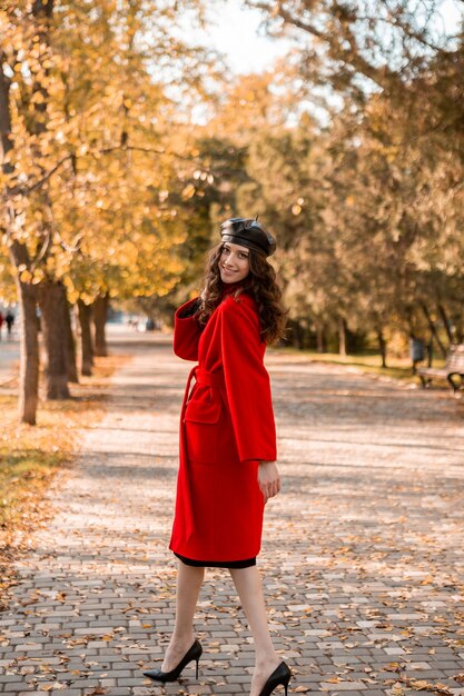 Grande jolie femme maigre souriante élégante avec des cheveux bouclés marchant dans le parc habillé en manteau rouge chaud automne mode tendance, style de rue, portant un béret