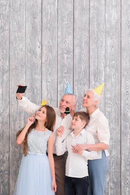 Grands-parents portant un chapeau de fête prenant un selfie sur un téléphone portable avec leurs petits-enfants tenant des accessoires en papier