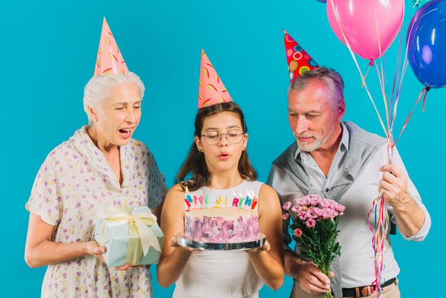 Grands-parents tenant des cadeaux d&#39;anniversaire près de fille avec gâteau soufflant des bougies sur fond bleu