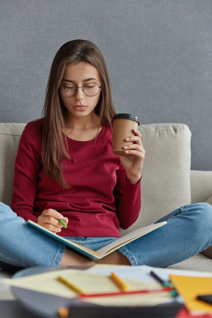 La graphiste féminine aux cheveux noirs songeuse fait la notation dans un cahier, écrit des informations, garde les jambes croisées, porte une tasse de café jetable