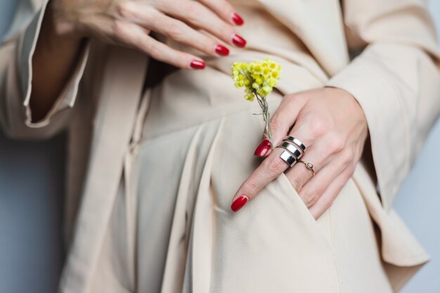 Gros coup de femme mains manucure rouge deux anneaux portant un costume beige. Jolie fleur séchée jaune dans la poche.