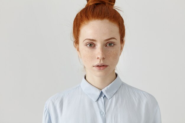 Gros coup de jolie jeune femme aux cheveux roux réussie en chemise formelle ayant un regard sérieux et confiant avant la journée de travail, posant au mur de bureau blanc. Concept de personnes et de style de vie