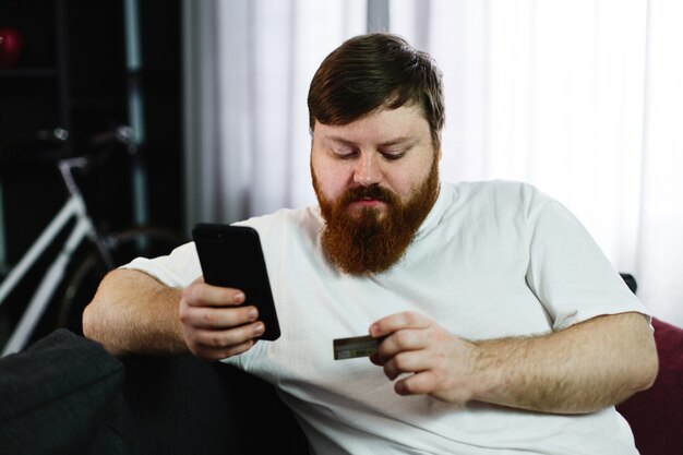 Gros homme tape le numéro d&#39;une carte de crédit dans son téléphone assis sur le canapé