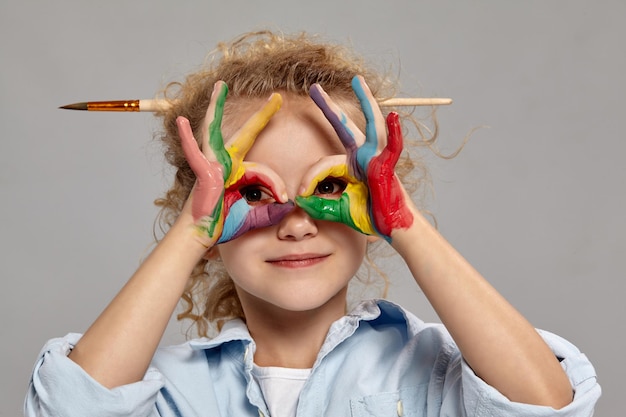 Gros plan d'un adolescent drôle ayant une brosse dans ses cheveux blonds bouclés chics, vêtu d'une chemise bleue et d'un t-shirt blanc. Elle a plié ses mains peintes comme si ce sont ses lunettes et souriant à la caméra