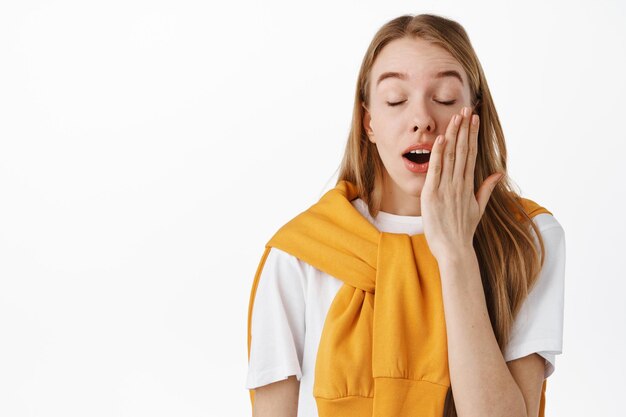 Gros plan d'une adolescente fatiguée ou ennuyée, bâillant les yeux fermés, couvrant la bouche ouverte, écoutant quelque chose d'ennuyeux, debout sur fond blanc