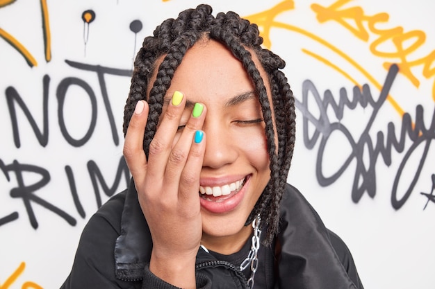 Photo gratuite gros plan d'une adolescente heureuse fait le visage sourit de paume a largement manucure colorée et dreadlocks exprime des émotions positives pose contre un mur de graffitis dessiné vêtu de vêtements à la mode