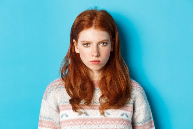 Gros plan d'une adolescente rousse en colère qui a l'air énervée contre la caméra, fronce les sourcils et boude en colère, debout sur fond bleu.