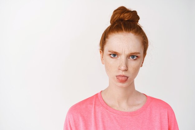 Photo gratuite gros plan d'une adolescente rousse idiote montrant la langue et fronçant les sourcils, debout contre le mur blanc