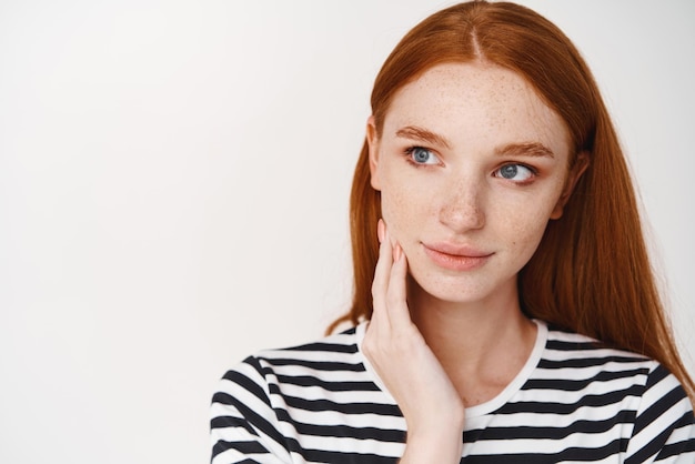 Gros plan d'une adolescente rousse avec des taches de rousseur et des yeux bleus regardant à gauche l'espace de copie touchant une peau propre et saine sur le visage debout sur fond blanc