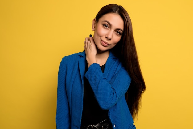 Gros plan adorable belle femme en veste bleue posant avec le sourire sur le mur jaune. Modèle féminin européen posant