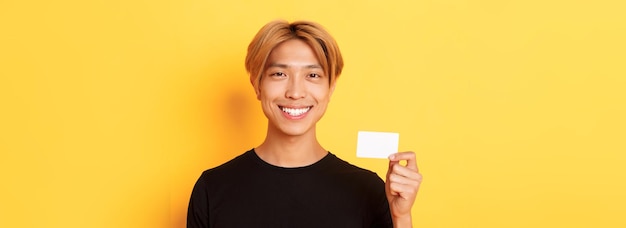 Gros plan d'un beau mec asiatique heureux avec des cheveux blonds souriant heureux et montrant une carte de crédit sur crier
