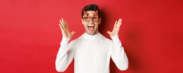 Gros plan d'un beau mec surpris en pull blanc et lunettes de fête entendre une incroyable promo de Noël de...