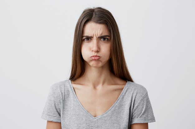 Gros plan de la belle charmante drôle jeune femme de race blanche aux cheveux longs noirs en chemise grise décontractée gonfler les joues, avec l'expression du visage insatisfait et offensé, s'amuser sur grad
